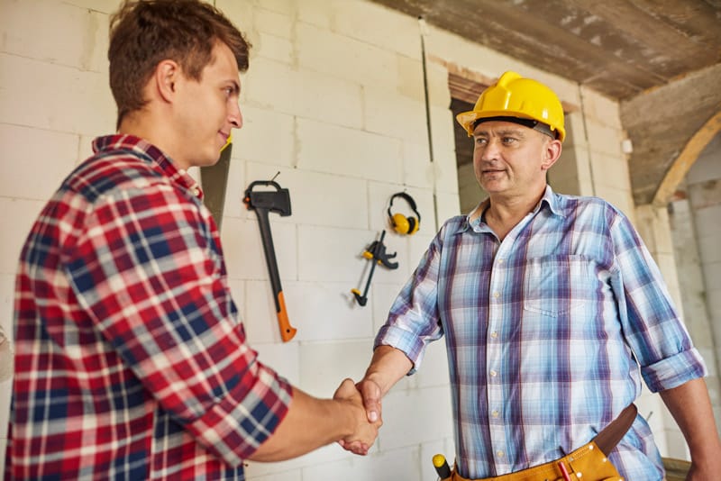 Un artisan en casque et ceinture à outils serre la main d'un client sur un chantier de rénovation